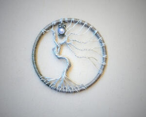 Handmade wire art shows a silver tree with twisted roots and branches inside a circular rustic frame, topped with a pearl-bodied owl charm.